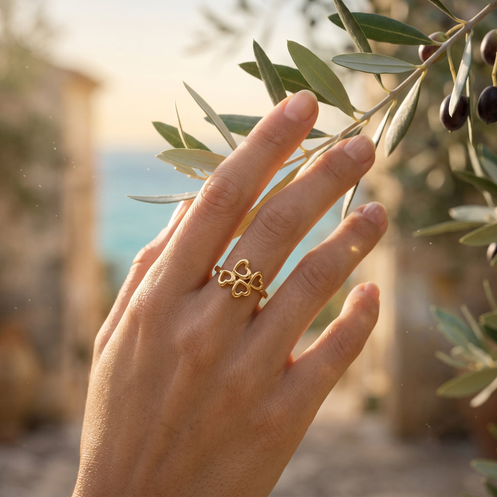 Macro editorial shot of the Lamar four hearts ring, 18k yellow gold, on a hand gently touching an olive branch.	لقطة مقربة لخاتم لامار بتصميم 4 قلوب، ذهب أصفر عيار 18، على يد تلمس غصن زيتون بلطف.