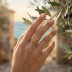 Macro editorial shot of the Lamar four hearts ring, 18k yellow gold, on a hand gently touching an olive branch.	لقطة مقربة لخاتم لامار بتصميم 4 قلوب، ذهب أصفر عيار 18، على يد تلمس غصن زيتون بلطف.