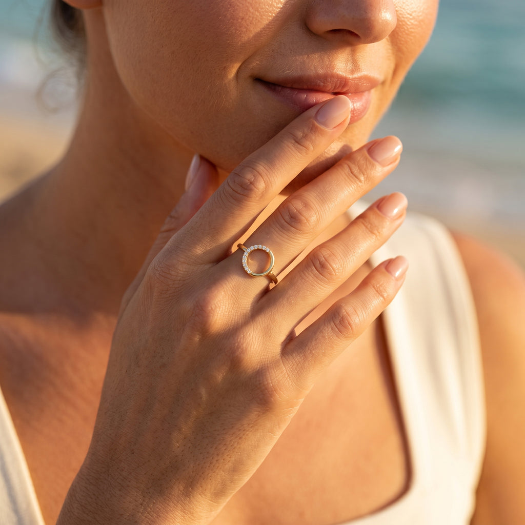 Feminine hand wearing the King Gold open circle ring, 18k gold, touching lips on a Mediterranean beach.	يد أنثوية ترتدي خاتم كينج جولد الدائري، ذهب عيار 18، بالقرب من الشفاه على شاطئ متوسطي.