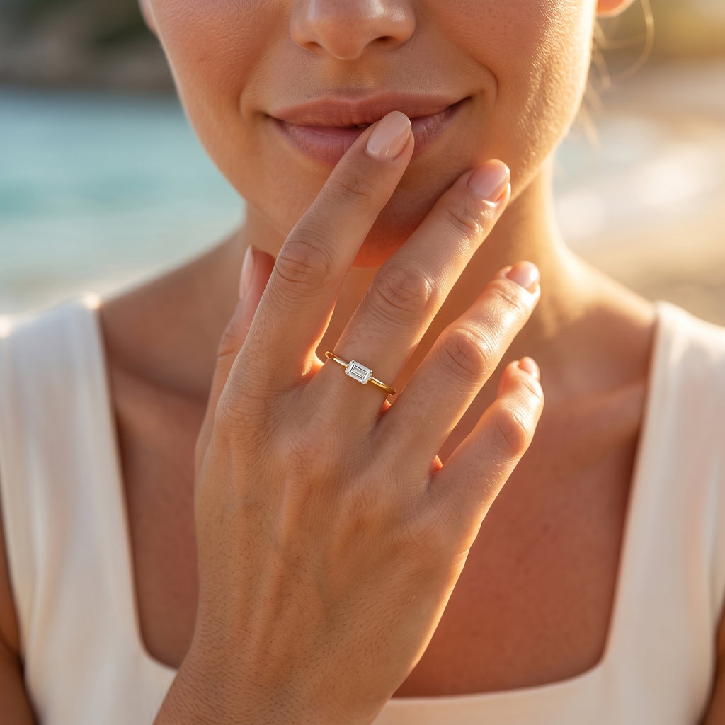 Feminine hand wearing the sleek Andria 18k gold white stone ring, touching lips with a Mediterranean beach background.	يد أنثوية ترتدي خاتم أندريا الرقيق من الذهب عيار 18 بفص أبيض، بالقرب من الشفاه مع خلفية شاطئ متوسطي.