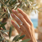 Close-up of the delicate Andria 18k gold ring on a hand gently touching a Mediterranean olive branch.	لقطة مقربة لخاتم أندريا الرقيق من الذهب عيار 18 على يد تلمس غصن زيتون متوسطي بلطف.