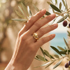 Close-up of a feminine hand touching an olive branch, wearing a delicate 4g L'azurde 18k gold scattered stone ring. لقطة مقربة ليد رقيقة تلمس غصن زيتون وترتدي خاتم لازوردي بومبيه ذهب عيار 18 بوزن 4 جرام.