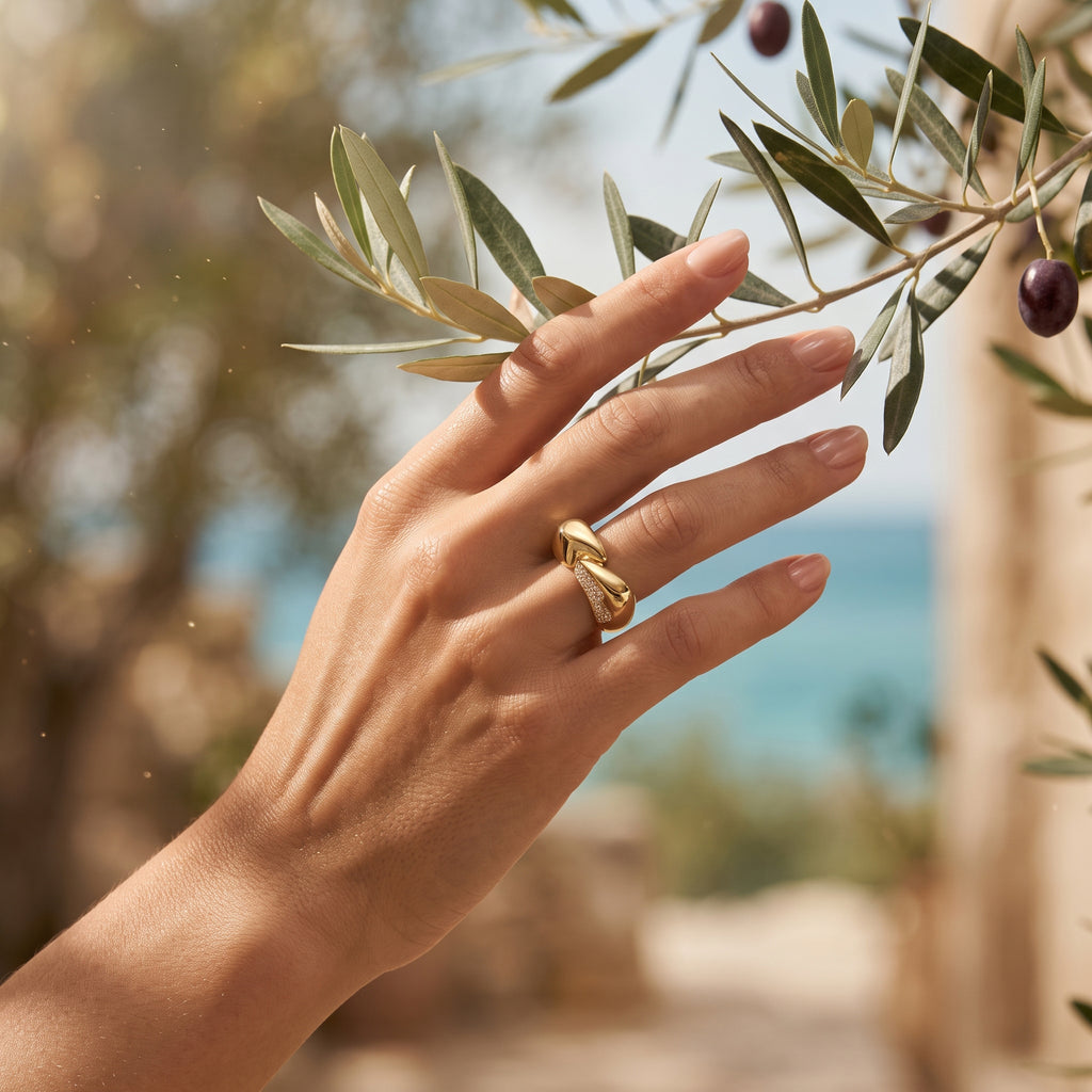 Feminine hand delicately touching an olive branch, wearing a 5.37g L'azurde 18k gold bypass ring. يد رقيقة تلمس غصن زيتون بلطف وترتدي خاتم لازوردي التفافي من الذهب عيار 18 بوزن 5.37 جرام.