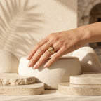 Elegant woman's hand wearing the Andria gold ring resting on a sculptural ivory pedestal.	يد امرأة أنيقة ترتدي خاتم أندريا الذهبي وتستند على قاعدة نحتية فاخرة.