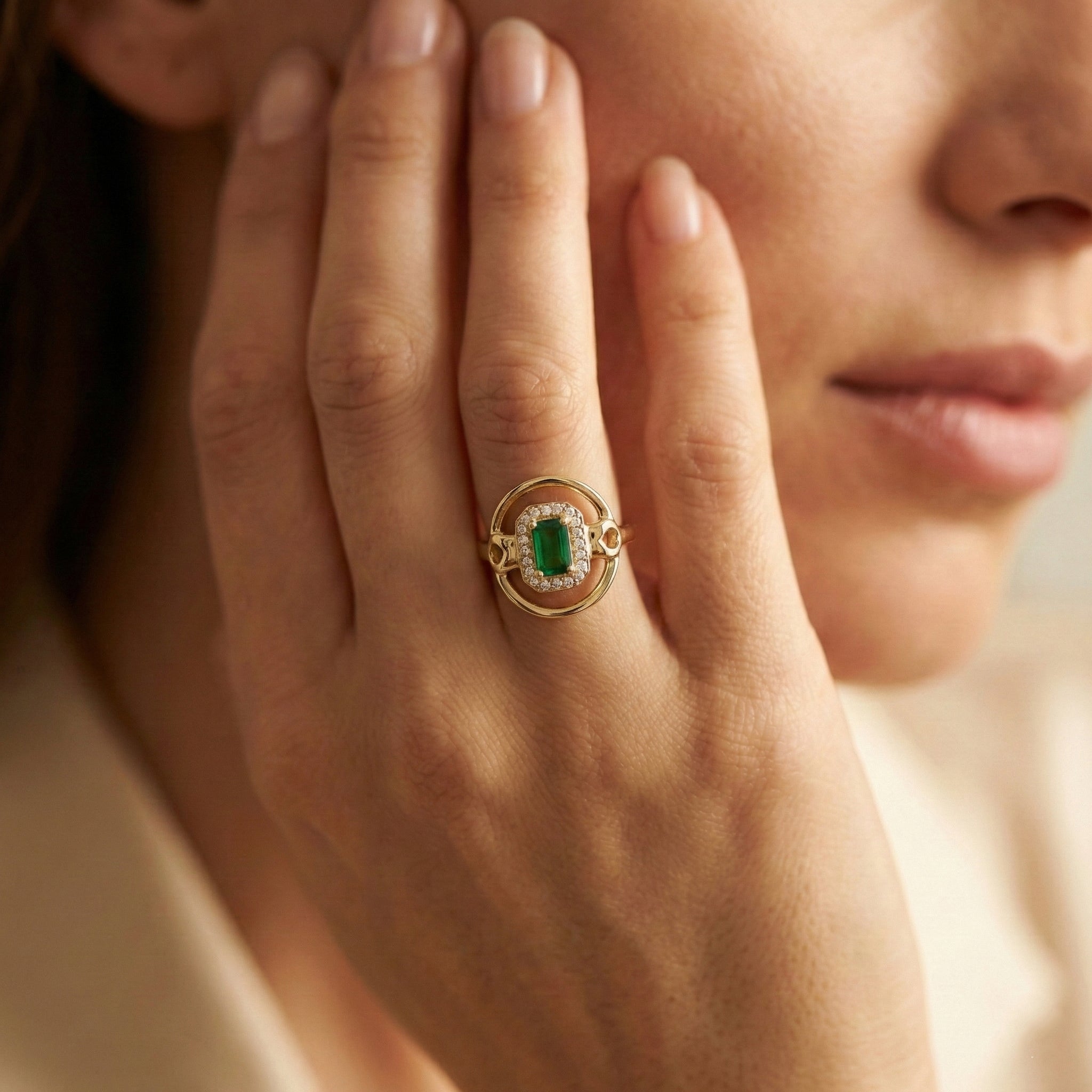 Close-up of a hand wearing a gold ring with a green gemstone, held near the face.