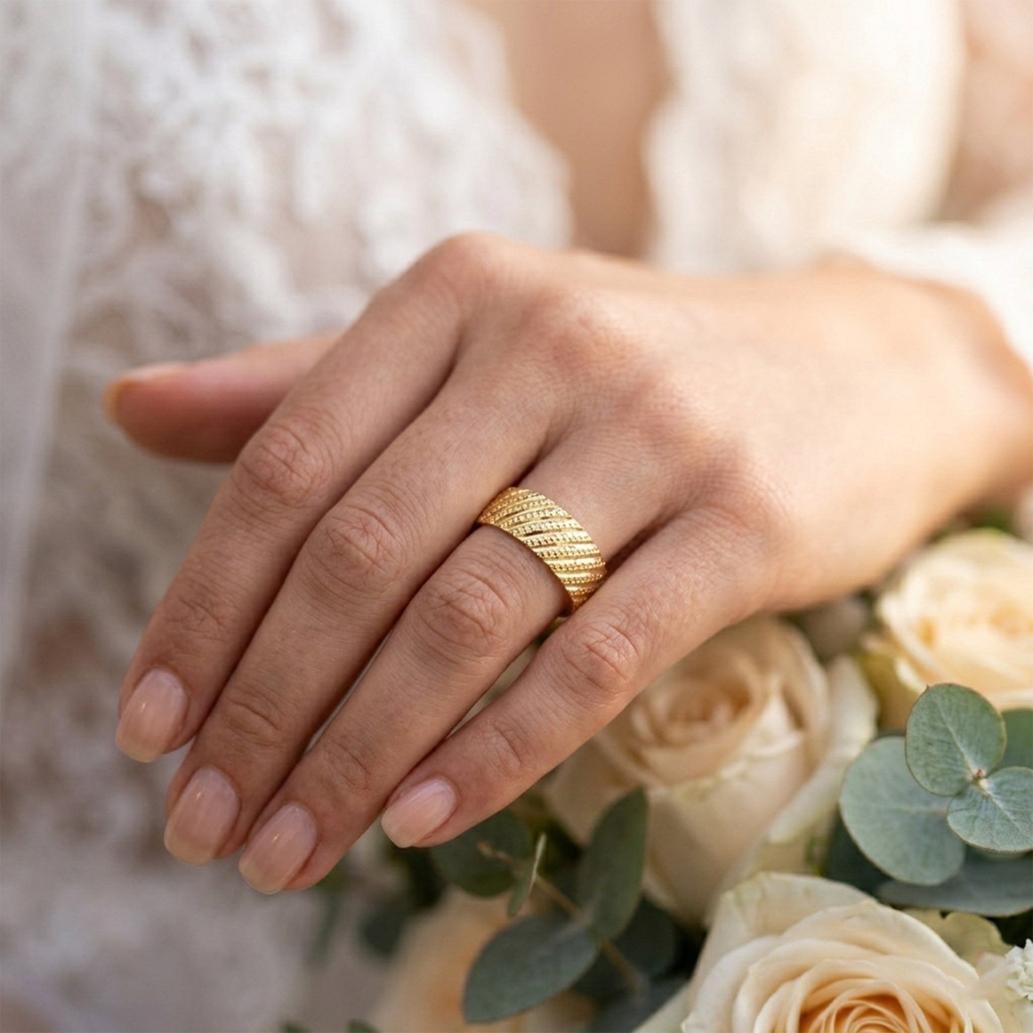 Alyans 18k yellow gold wedding band ring from The Forever Edit collection, elegantly worn on a woman's ring finger. The hand rests gracefully against a romantic, soft-focus bridal background of cream roses, delicate white lace, and eucalyptus leaves.