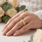 Close-up of a woman's hand wearing the Alyans 18k yellow gold wedding band ring from The Forever Edit category, resting softly against a blurred, romantic background of white lace and cream roses.
